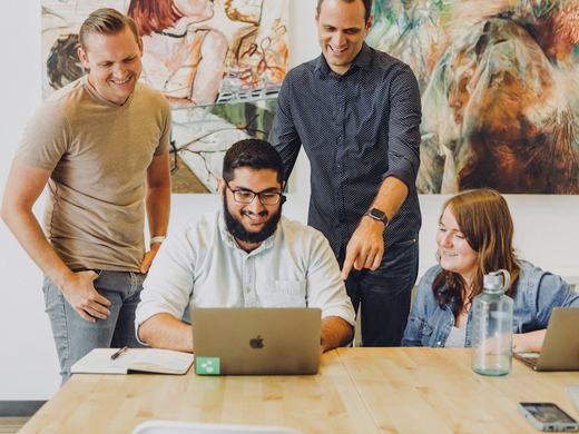 Grupo de pessoas sorrindo em volta de um notebook em uma mesa de trabalho